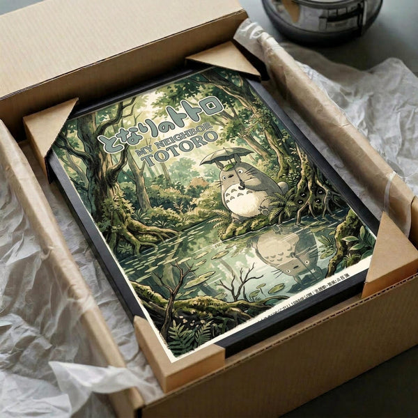 May include: Framed Studio Ghibli My Neighbor Totoro poster featuring the iconic forest spirit character sitting by a woodland stream, surrounded by lush green trees and foliage in a dreamy, illustrated art style. The vintage-toned print shows Japanese text at the top and is displayed in a black frame, carefully packaged in brown cardboard with protective wrapping materials on a white surface.
