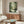 May include: Studio Ghibli My Neighbor Totoro movie poster displayed above wooden mid-century modern credenza in minimalist living room. Forest scene shows gray Totoro character with umbrella reflected in water, surrounded by lush green trees. Cream upholstered chair, glass coffee table, and modern lamp complete the neutral-toned space with natural lighting.