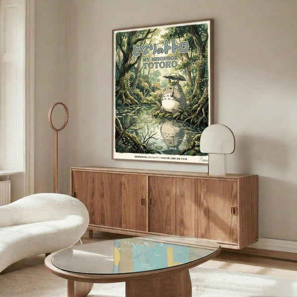 May include: Studio Ghibli My Neighbor Totoro movie poster displayed above wooden mid-century modern credenza in minimalist living room. Forest scene shows gray Totoro character with umbrella reflected in water, surrounded by lush green trees. Cream upholstered chair, glass coffee table, and modern lamp complete the neutral-toned space with natural lighting.