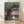 May include: Framed Studio Ghibli poster print leaning against white wall on light wood floor. Features My Neighbor Totoro artwork with large tree, cat bus in branches with glowing windows, two children and Totoro characters beneath. Soft twilight colors in purple, green, and cream. Japanese text visible. Natural wood frame. Warm lighting creates gentle shadows on textured wall.