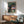 May include: Studio Ghibli My Neighbor Totoro movie poster displayed in modern living room with mid-century wooden credenza, sage green upholstered chair, and ceramic table lamp. Poster shows iconic Totoro characters under large tree with Catbus above, rendered in warm earth tones with green and purple sky. Natural sunlight streams across white walls creating geometric shadows.
