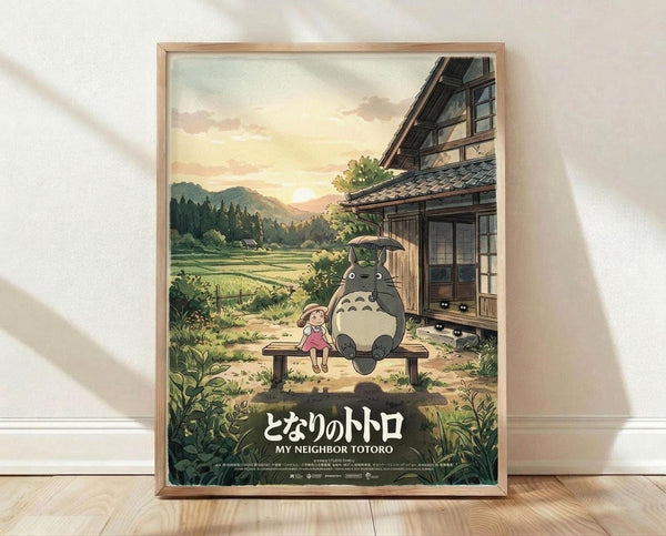 May include: Framed poster displaying animated scene with large gray forest creature and small child sitting together on wooden bench beside traditional Japanese house. Warm sunset sky with mountains and green fields in background. Natural wood frame leaning against white textured wall on light wooden floor with soft window shadows.