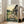 May include: Framed Studio Ghibli movie poster showing Totoro and young girl sitting on wooden bench outside traditional Japanese house at sunset. Warm golden sky with rolling green hills and rice fields in background. Poster leans against beige sofa in bright white living room with large windows and wooden floors.