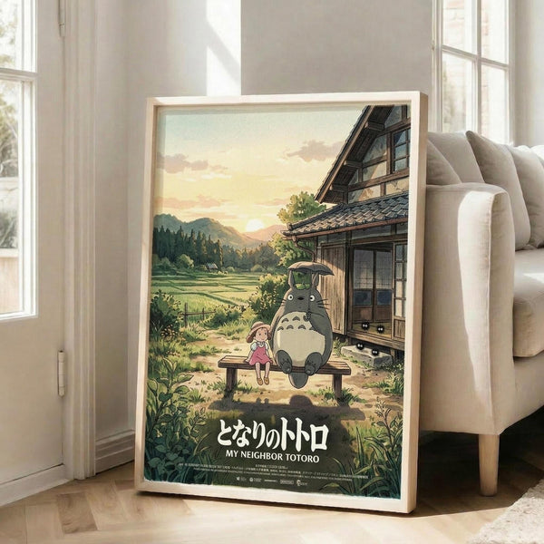 May include: Framed Studio Ghibli movie poster showing Totoro and young girl sitting on wooden bench outside traditional Japanese house at sunset. Warm golden sky with rolling green hills and rice fields in background. Poster leans against beige sofa in bright white living room with large windows and wooden floors.
