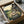 May include: Framed My Neighbor Totoro poster showing the iconic gray forest spirit and young girl Mei sitting together on wooden steps outside a traditional Japanese house. The artwork features warm sunset colors with rolling green hills and countryside in the background. The black-framed print is carefully packed in brown cardboard shipping box with white tissue paper padding, photographed on white fabric surface.