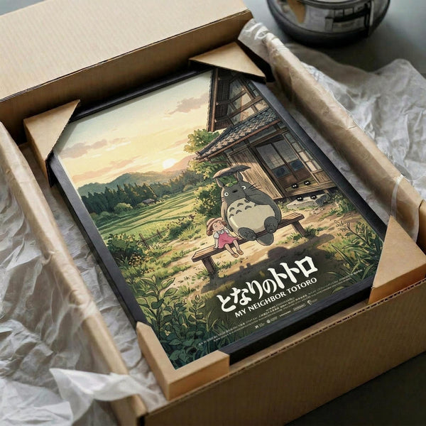 May include: Framed My Neighbor Totoro poster showing the iconic gray forest spirit and young girl Mei sitting together on wooden steps outside a traditional Japanese house. The artwork features warm sunset colors with rolling green hills and countryside in the background. The black-framed print is carefully packed in brown cardboard shipping box with white tissue paper padding, photographed on white fabric surface.