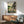 May include: Framed Studio Ghibli poster featuring Totoro and small child sitting on wooden bench outside traditional Japanese house, surrounded by green countryside and mountains at sunset. Wood-framed print hangs above mid-century modern credenza with white ceramic lamp, next to sage green upholstered armchair with wooden arms in bright, minimalist living room with white walls and natural lighting.