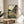 May include: Framed Studio Ghibli movie poster displayed on round wooden side table next to beige upholstered chair. Artwork shows iconic gray Totoro character sitting on wooden bench with young girl under umbrella, featuring lush green countryside landscape with traditional Japanese house, mountains, and golden sunset sky. Natural wood frame complements warm, sunlit room setting with plant leaves visible in corner.