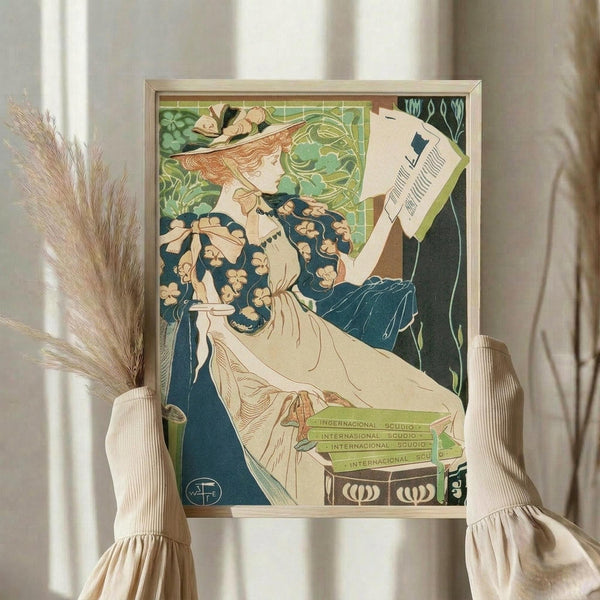 May include: Framed vintage Art Nouveau poster showing elegant woman in flowing white dress and floral blue shawl reading papers, with decorative green foliage background. Light wood frame displayed on white ribbed fabric chair with dried pampas grass beside it in soft natural lighting.