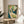 May include: Art Nouveau poster print in wooden frame displayed on round wooden side table next to beige upholstered chair. Artwork features elegant woman with flowing auburn hair in white dress against green background with ornate decorative elements. Soft natural lighting creates shadows on neutral wall. Plant leaves visible in corner of minimalist interior setting.