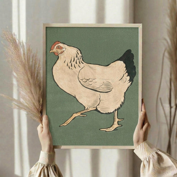 May include: Person in cream ruffled sleeves holding framed vintage Japanese woodblock print of white hen with red comb and black tail feathers on sage green background in light wood frame, with dried wheat stems visible to left, soft natural lighting from nearby window