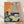May include: Framed art print leaning against white wall on light wood floor. The artwork shows a colorful still life with Asian-inspired elements - a green dragon against pink and orange backgrounds in the upper portion, and various bowls, cups, and yellow fruit arranged on a dark blue table surface below. The print has a vintage, hand-printed appearance with visible texture and weathering. Natural light creates soft shadows on the white fabric backdrop.