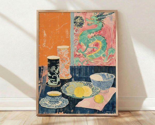 May include: Framed art print leaning against white wall on light wood floor. The artwork shows a colorful still life with Asian-inspired elements - a green dragon against pink and orange backgrounds in the upper portion, and various bowls, cups, and yellow fruit arranged on a dark blue table surface below. The print has a vintage, hand-printed appearance with visible texture and weathering. Natural light creates soft shadows on the white fabric backdrop.