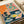 Colorful woodblock-style print featuring a still life scene with blue and white patterned teacups and bowls containing yellow citrus fruits on a pink tablecloth. Above hangs a vibrant coral and pink tapestry or screen decorated with an ornate green dragon design and medallion motif. The composition includes bold orange geometric shapes and rich navy blue areas, creating a striking contrast of warm and cool tones in an Asian-inspired artistic style.
