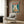 May include: Vintage-style poster featuring a white cat with golden eyes eating ramen noodles from a bowl with eggs and meat, displayed above a beige upholstered armchair with geometric pillow in a modern room with wood paneling, brass floor lamp, and side table with open book