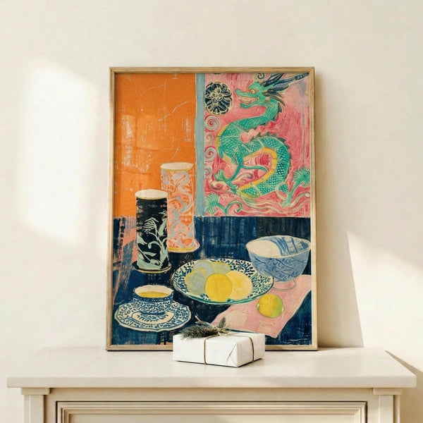 May include: Framed colorful still life artwork featuring a green dragon against pink background in upper section, with decorative bowls, yellow lemons, and ornate vessels arranged on a table below. The print sits on a light wooden console table against a cream wall, with a small white wrapped gift box positioned in front.