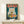 May include: Framed poster featuring a white cat with yellow eyes eating ramen noodles from a blue bowl, with red Japanese characters above on teal background. Wood frame displayed on light wooden shelf next to small white wrapped gift. Vintage-style illustration with detailed noodles, soft-boiled eggs, and meat toppings.
