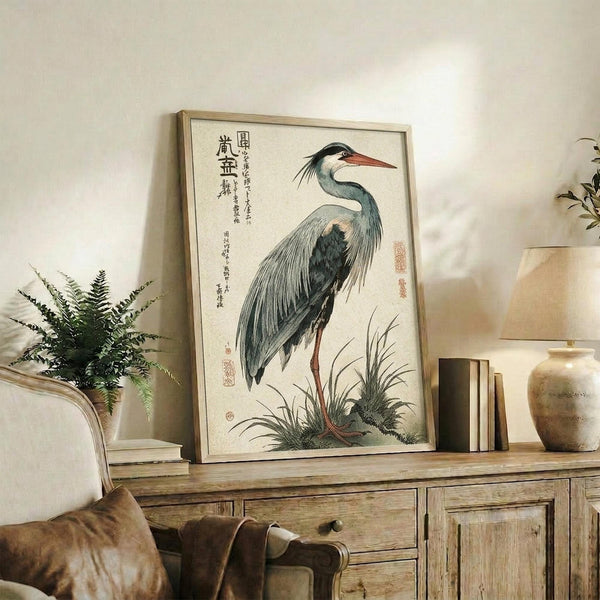 May include: Japanese woodblock print of a grey heron with long orange beak standing among marsh grasses, displayed in light wood frame on rustic wooden sideboard alongside fern plant, books, and ceramic vase in neutral-toned living room setting