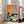 May include: A large framed art print leaning against a white wall next to a beige sofa in a bright living room. The artwork features a vibrant still life with Asian-inspired elements including a green dragon on coral pink background, blue ceramic bowls with yellow lemons, decorative vases, and a teacup on saucer. The composition uses bold colors of orange, pink, blue and yellow in a folk art printmaking style with visible texture and weathered edges.