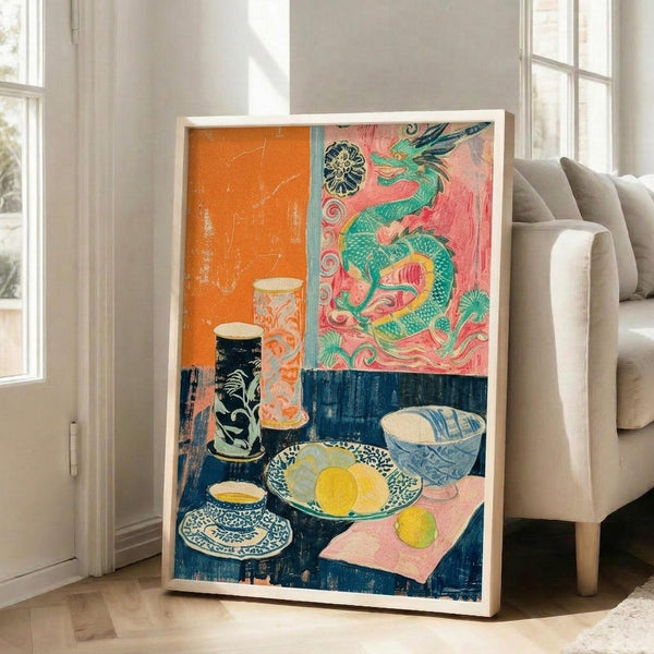 May include: A large framed art print leaning against a white wall next to a beige sofa in a bright living room. The artwork features a vibrant still life with Asian-inspired elements including a green dragon on coral pink background, blue ceramic bowls with yellow lemons, decorative vases, and a teacup on saucer. The composition uses bold colors of orange, pink, blue and yellow in a folk art printmaking style with visible texture and weathered edges.