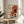 May include: Japanese-style art print in wooden frame showing red octopus with flowing tentacles against cream background, featuring blue ocean wave and large red sun. Print displayed on round wooden side table next to beige upholstered chair with white pillow. Soft natural lighting creates shadows on neutral wall, with plant leaves visible in corner.