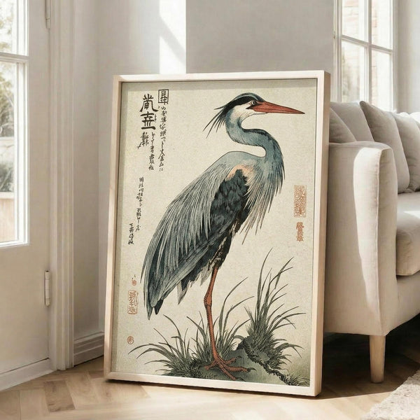 May include: A framed Japanese woodblock print featuring a grey heron with blue-grey plumage and orange beak standing among marsh grasses. The artwork displays traditional ukiyo-e style with Japanese calligraphy and red seals on cream paper. The print is shown in a light wood frame leaning against white walls in a bright living room with beige furniture and large windows.