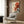 May include: Japanese-style artwork showing a red octopus with curling tentacles against a cream background with orange sun and blue ocean wave, displayed in light wood frame on wood-paneled wall above beige armchair with geometric pillow, brass floor lamp and dark side table with open book nearby