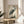 May include: Japanese woodblock print of a grey heron with long orange beak and dark wings standing among marsh grasses, displayed in thin wooden frame on round wooden side table next to beige upholstered chair. Natural sunlight streams across white wall creating soft shadows from nearby plant leaves.