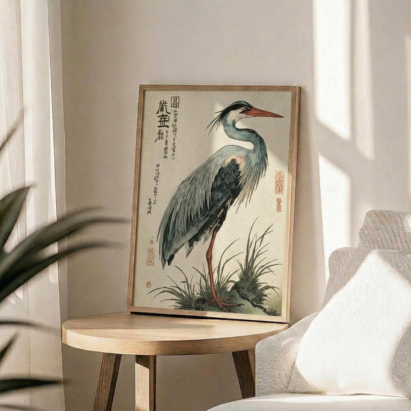 May include: Japanese woodblock print of a grey heron with long orange beak and dark wings standing among marsh grasses, displayed in thin wooden frame on round wooden side table next to beige upholstered chair. Natural sunlight streams across white wall creating soft shadows from nearby plant leaves.