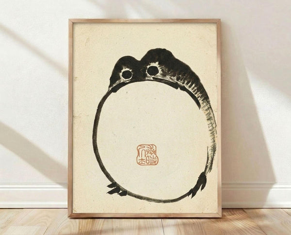 May include: Framed Japanese ink painting of a minimalist black frog curled in a circular pose on cream textured paper, displayed in light wood frame against white wall on wooden floor. The artwork features bold brushstrokes in traditional sumi-e style with a red square seal stamp, creating an elegant zen-like composition with natural lighting casting soft shadows.