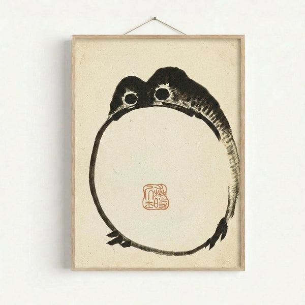May include: Framed Japanese ink painting hanging on white wall showing minimalist black brushstroke frog in circular pose with two round eyes and webbed feet, red square seal stamp in center, displayed in light wood frame with hanging cord against cream textured paper background
