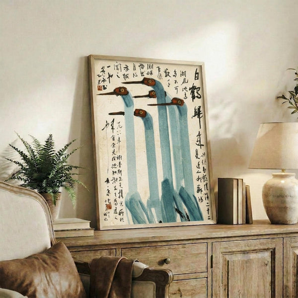 May include: Framed Asian-style painting featuring five tall blue cranes with brown heads arranged in a line, accompanied by black Chinese calligraphy on white background. Light wood frame displayed on rustic wooden credenza alongside fern plant, ceramic vase, and books in neutral-toned living room setting.