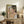 May include: Framed poster print of expressionist figure painting featuring intertwined nude forms in muted pink, green and earth tones with bold black outlines, displayed on rustic wooden sideboard alongside fern plant, ceramic vase, and books in neutral living room setting with beige chair visible in foreground