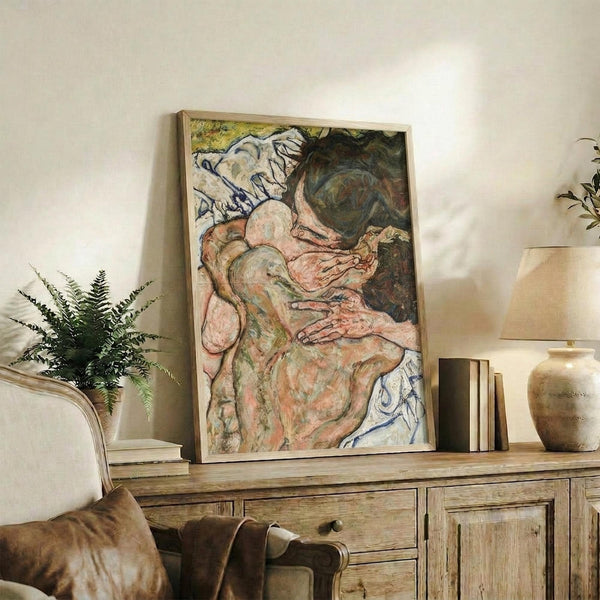 May include: Framed poster print of expressionist figure painting featuring intertwined nude forms in muted pink, green and earth tones with bold black outlines, displayed on rustic wooden sideboard alongside fern plant, ceramic vase, and books in neutral living room setting with beige chair visible in foreground