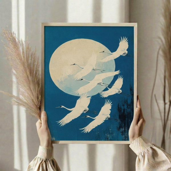 May include: Person in cream ruffled sleeves holding framed vintage Japanese art print featuring white cranes flying across deep blue sky with large pale moon. Print shows traditional woodblock style with textural brushwork at bottom. Person also holds dried pampas grass stems. Soft neutral curtains and lighting in background create serene styling scene.
