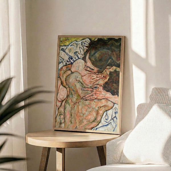 May include: Framed poster print of an expressionist painting showing two embracing figures in muted tones of pink, green, and blue, displayed on a round wooden side table next to a beige upholstered chair. Natural sunlight streams across the neutral-toned room setting, with a plant visible in the corner. The artwork features bold, emotional brushstrokes and distorted forms typical of early 20th century expressionist style.
