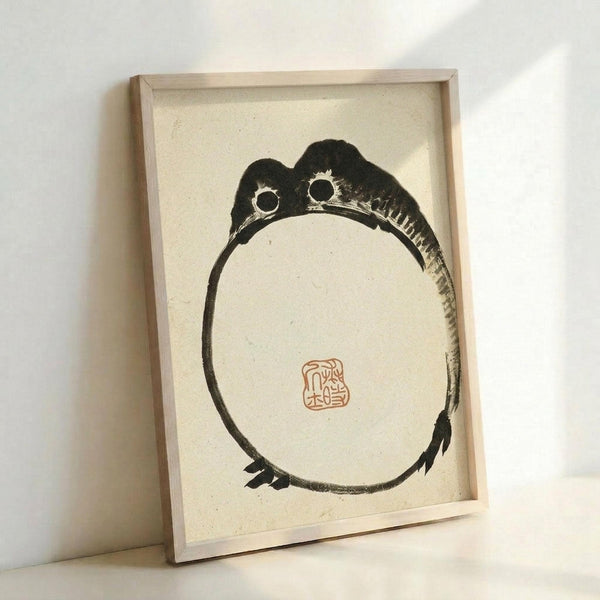 May include: Framed artwork featuring a minimalist black ink drawing of a round frog with large bulging eyes and webbed feet, painted in traditional Japanese brush style on cream textured paper. A small red rectangular seal stamp appears in the center. The piece is displayed in a simple light wood frame, leaning against a white wall with soft natural lighting creating gentle shadows.