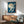May include: Framed Japanese-style print featuring white cranes flying against deep blue night sky with large pale moon, displayed above mid-century modern wood credenza with table lamp, next to sage green upholstered armchair in sunlit room with white walls