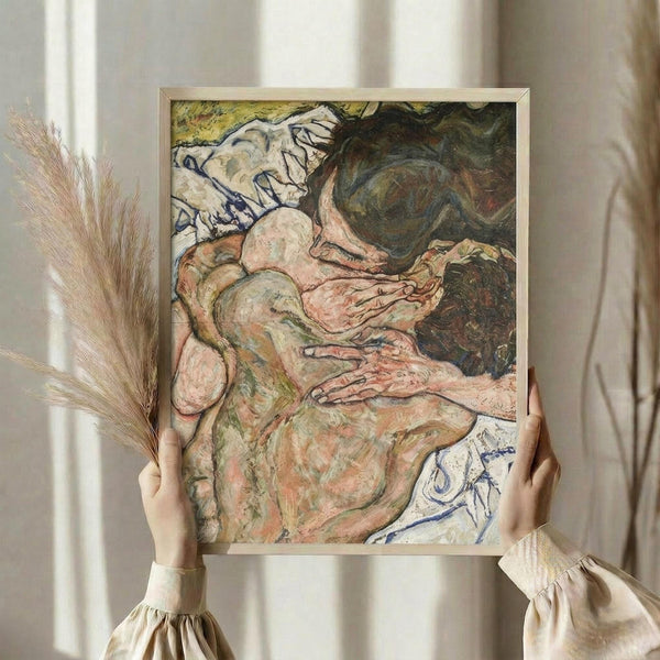 May include: Person in cream sweater holding framed art print showing expressionist painting of two embracing nude figures in earthy tones of pink, green and brown with blue accents. Dried pampas grass visible to left, soft natural lighting on white curtained background.