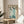 May include: Framed Asian-style artwork featuring five stylized cranes with blue-gray necks, brown heads, and long dark beaks, surrounded by Chinese calligraphy text on cream paper. The thin wooden frame sits on a round light wood side table next to a beige upholstered chair, with plant leaves visible in soft natural lighting against white walls.