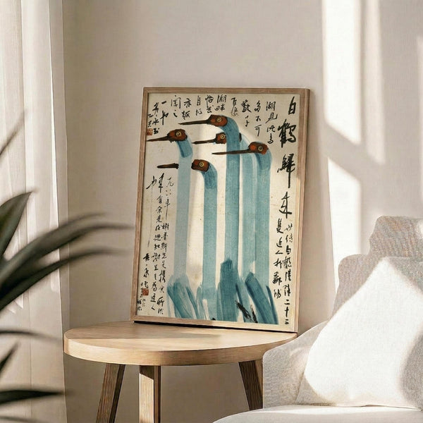 May include: Framed Asian-style artwork featuring five stylized cranes with blue-gray necks, brown heads, and long dark beaks, surrounded by Chinese calligraphy text on cream paper. The thin wooden frame sits on a round light wood side table next to a beige upholstered chair, with plant leaves visible in soft natural lighting against white walls.