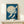 May include: Framed vintage Japanese woodblock print showing white cranes flying across deep blue night sky with large pale moon, displayed on light wood console table with small wrapped gift box, against cream wall with natural lighting