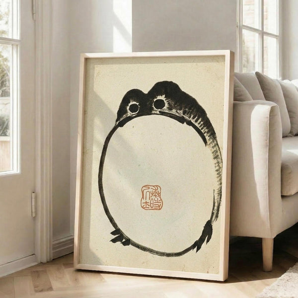 May include: Large framed artwork leaning against white wall next to beige upholstered chair in bright room with tall windows. The print features a minimalist black ink painting of a round frog with prominent eyes on cream-colored textured paper, with a small red traditional seal stamp. Natural wood frame, sunlight streaming across wooden floors.