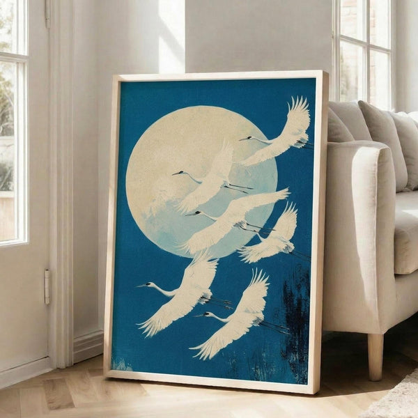 May include: A framed vintage Japanese-style poster featuring white cranes flying against a deep blue sky with a large pale yellow moon. The artwork shows multiple elegant birds with outstretched wings in different flight positions, rendered in soft cream and blue tones. The print is displayed in a light wooden frame, leaning against white walls near tall windows in a bright living room with a beige upholstered chair visible.