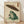 May include: Framed Japanese woodblock style print featuring a green frog holding a traditional paper umbrella in falling rain. The artwork displays muted earth tones with detailed cross-hatching technique and Japanese characters in red cartouches. Light wood frame sits on wooden floor against white wall with natural lighting creating soft shadows.