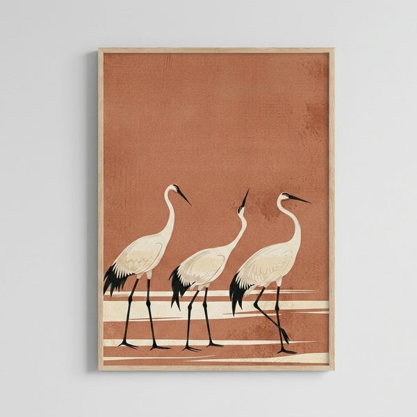 May include: Framed art print displayed on white wall showing three white cranes with black wing tips and long dark beaks standing in a row against a warm terracotta orange background with horizontal cream and brown striped bands. The birds have elegant long necks and legs in a minimalist Japanese-inspired artistic style within a light wooden frame.