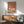 May include: Framed poster with three white cranes on warm terracotta background displayed above mid-century modern wood credenza. Scene shows sage green upholstered armchair with wooden arms, white ceramic table lamp, and natural sunlight casting shadows across white wall. Artwork features minimalist Japanese-inspired design with elegant long-necked birds standing on geometric striped ground in cream and brown tones.