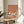 May include: Framed poster showing three white cranes with black wing tips and long beaks standing on coral-orange background, displayed on round wooden side table next to beige upholstered chair with white throw pillow, green plant leaves visible, natural sunlight casting shadows on cream wall