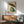 May include: Japanese woodblock style print featuring a green frog sitting under a brown paper umbrella in falling rain, displayed in a wooden frame above a mid-century modern credenza with a small white table lamp. The artwork shows traditional Asian art styling with muted earth tones against a textured background. A sage green upholstered armchair with wooden arms sits nearby in the bright, minimalist room setting.