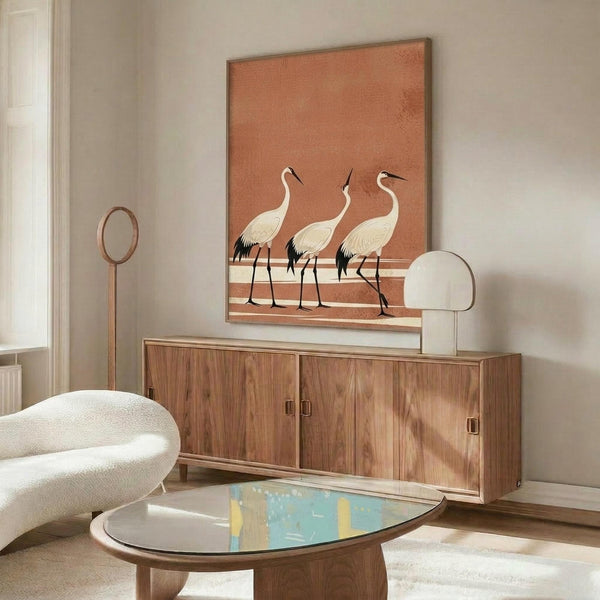 May include: Japanese-inspired artwork featuring three elegant white cranes with black wing tips and long necks, set against a warm terracotta orange background with horizontal bands at the bottom. The framed poster hangs above a mid-century modern walnut credenza in a minimalist living room with cream walls, white upholstered chair, glass-top coffee table, and contemporary lighting fixtures.